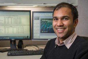 a man, smiling and looking into the camera with computer monitors behind him.